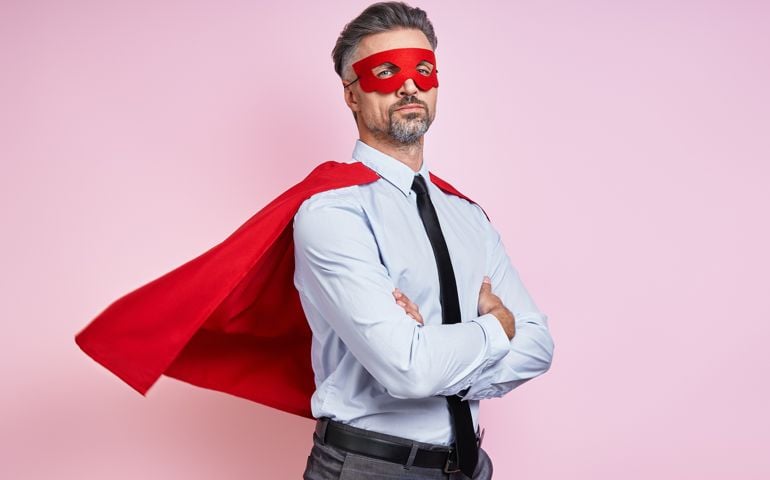 Confident man in shirt and tie wearing superhero cape and keeping arms crossed against pink background
