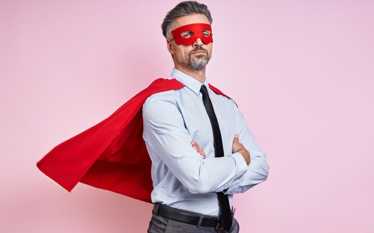Confident man in shirt and tie wearing superhero cape and keeping arms crossed against pink background