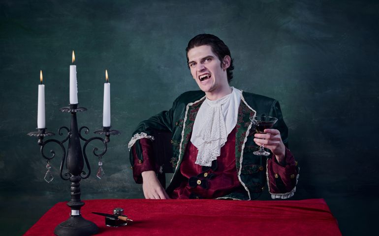Portrait of terrifying man in image of vampire, Dracula rising glass of blood over dark green background. Grinning