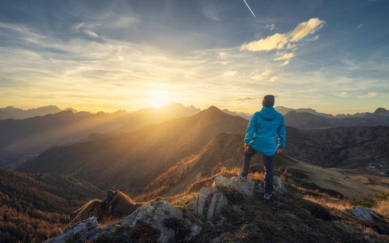 Man on stone on the hill and beautiful mountains in haze at colorful sunset in autumn. Dolomites, Italy. Sporty guy, mountain ridges in fog, orange grass and trees, blue sky with sun in fall. Hiking