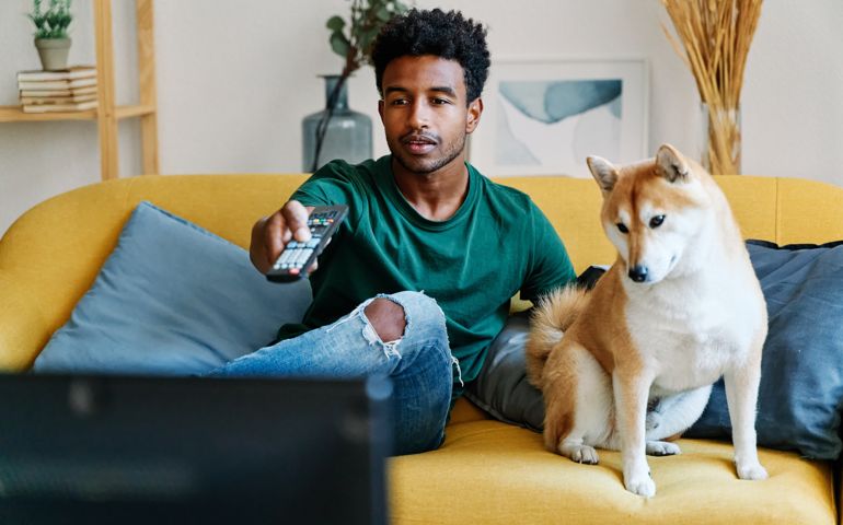 Man sitting on sofa in living room with dog watching TV changing channels with remote control.