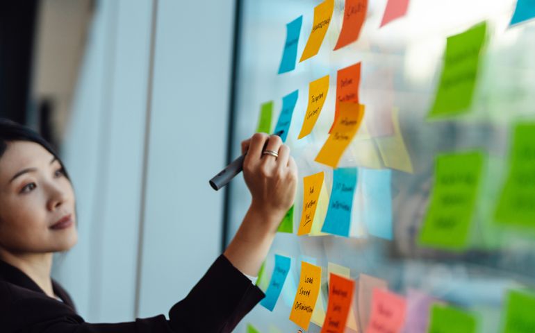 Young Asian businesswoman brainstorming strategy and new ideas on glass wall with adhesive notes