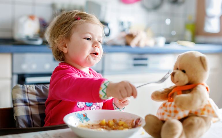 Adorable baby girl eating from fork vegetables and pasta. food, child, feeding and development concept. Cute toddler, daughter with spoon sitting in highchair and learning to eat by itself.
