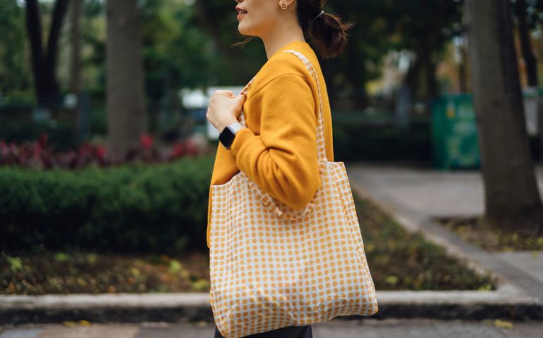 Side profile, cropped shot of young Asian woman walking in the city against trees outdoors, carrying a reusable shopping bag after shopping. Responsible shopping, zero waste, eco-friendly concept. Sustainable living lifestyle