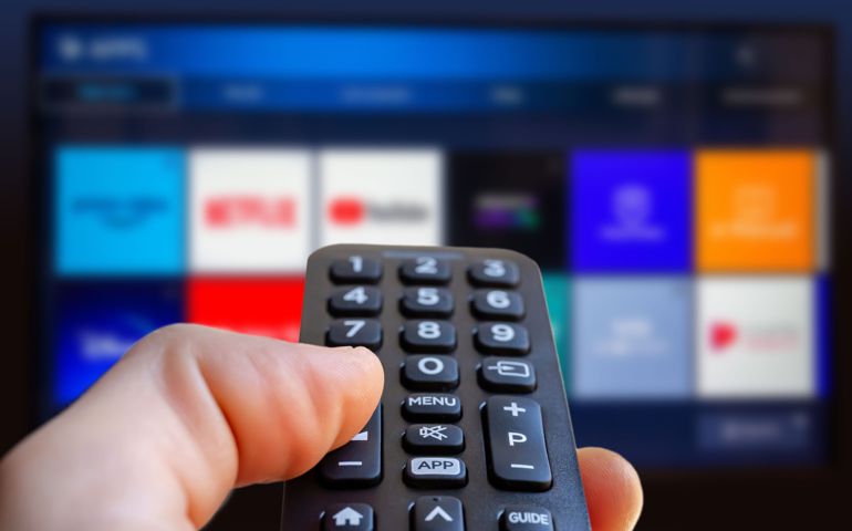 A man is holding a remote control of a smart TV in his hand. In the background you can see the television screen with streaming entertainment apps for video on demand