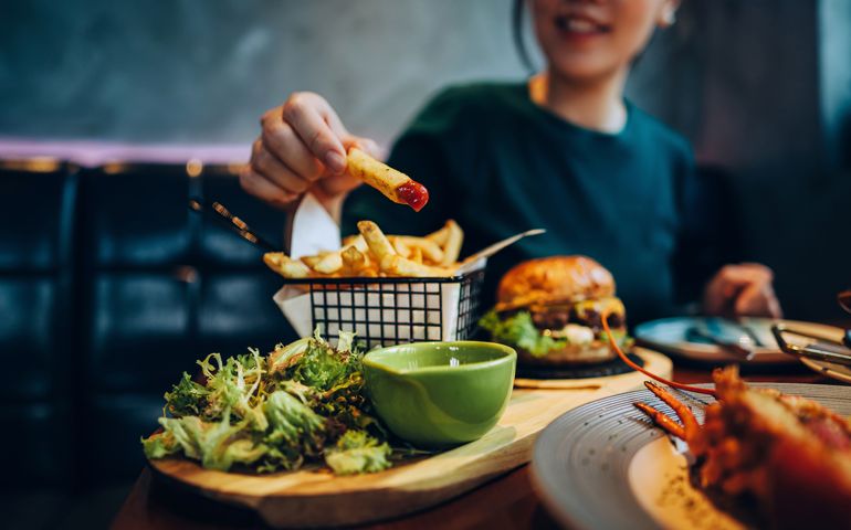 Cropped shot of young Asian woman eating freshly made delicious burger, dipping fries in ketchup on the dining table. Enjoying a feast in the restaurant. People, food and lifestyle