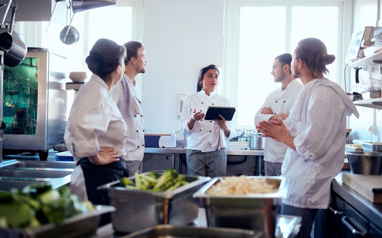 Mid adult female chef holding digital tablet while discussing with team in kitchen