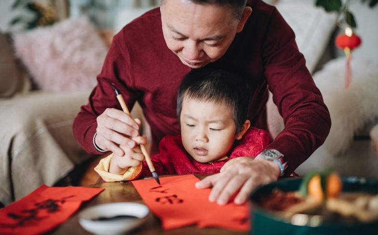 Grandfather practising Chinese calligraphy for Chinese New Year Fai Chun (Auspicious Messages) and teaching his grandson by writing it on a piece of red paper