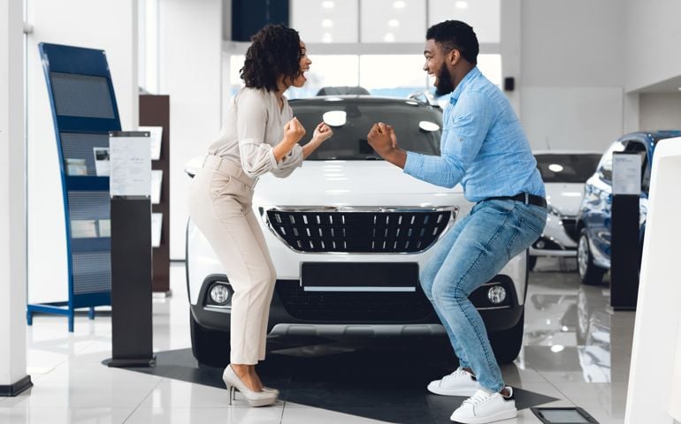 Excited Spouses Shaking Fists Standing Near New Auto In Dealership