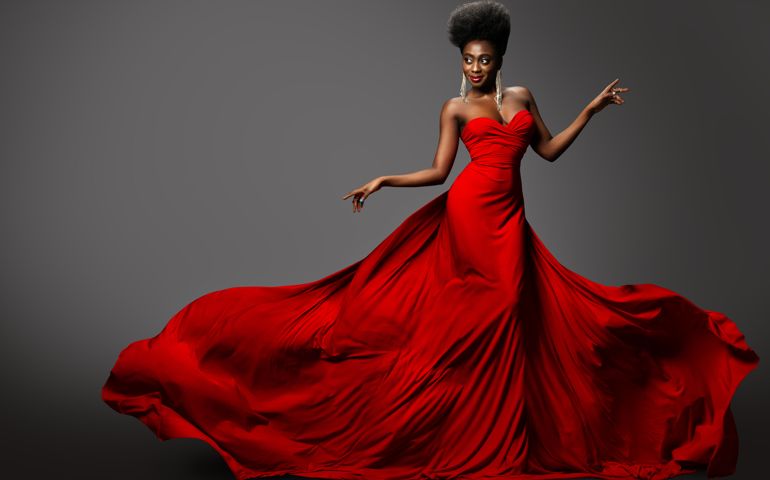 Fashion Model in Red Long waving Luxury Dress. Dark Skinned Beauty Woman with Afro Black Hairstyle dancing over Gray Background. Happy African Sexy Girl in Silk Evening Gown with flying Fabric on Wind