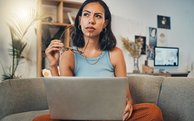 Beautiful mixed race woman thinking while using laptop for blogging in living room at home. Hispanic entrepreneur sitting cross legged alone on lounge sofa and planning next blog post on technology