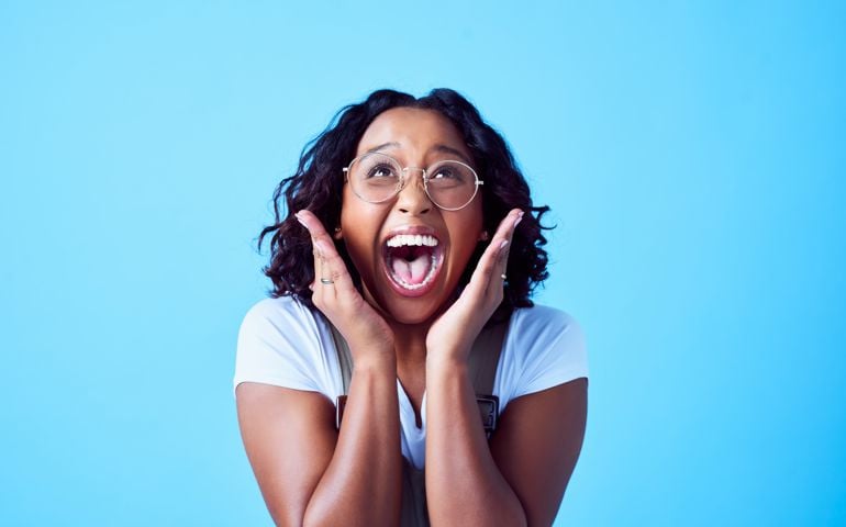 Excited, happy and surprised young female is amazed while looking up. Delight on a black or African woman's face against a bright blue background. Trendy and stylish lady celebrating being a winner