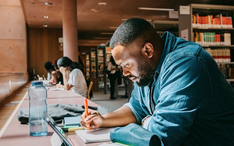 Student writing in notebook while sitting with tablet PC in college library