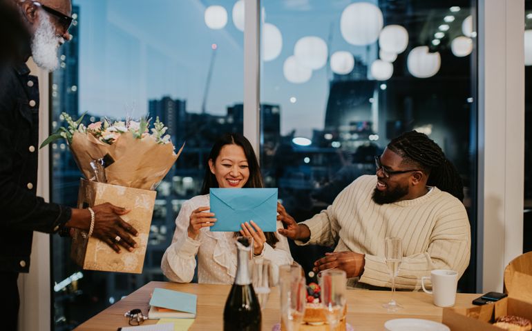 Office workers lavish attention and praise on a colleague, presenting a card and flowers to her as she smiles