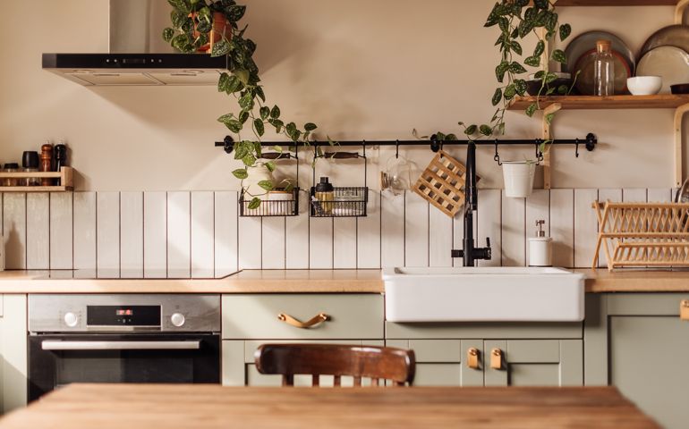 Empty kitchen island with marble surface in foreground, green vintage countertop with drawers and pendant lights hanging above, lots of flowers in jars, blurred background