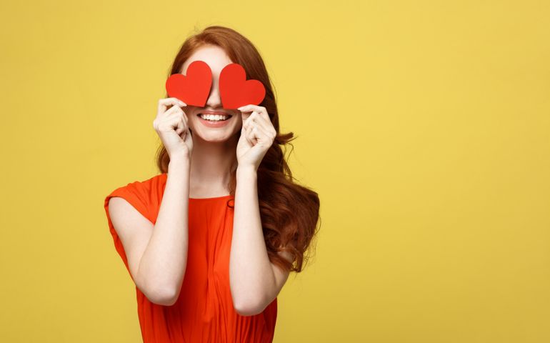 Lifestyle Concept: Attractive woman with beaming smile having two small red hearts in hands, closing eyes with paper heart symbols while standing over yellow background.