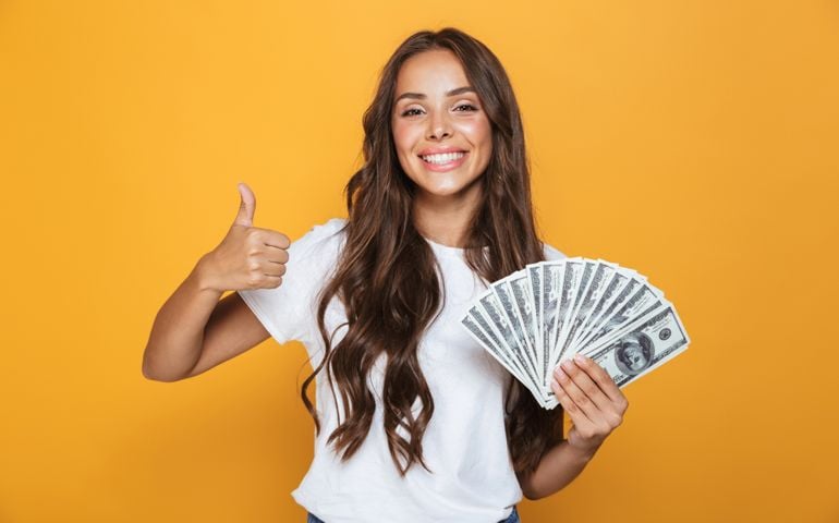 Portrait of a happy young girl with long brunette hair standing over yellow background, holding money banknotes, showing thumbs up