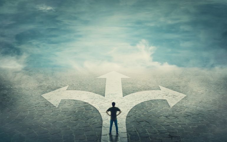 Confident man, hands on hips, stands in front of a crossroad with road split in three different ways as arrows. Choosing the correct pathway between left, right and front. Difficult decision concept.