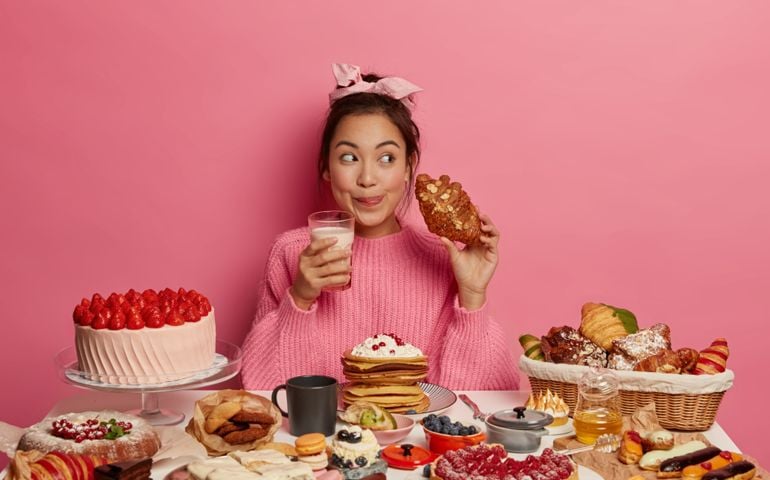 Addicted young Asian female drinks milk and eats delicious croissant, licks lips from pleasant taste, enjoys desirable favourite dessert, sits at table with many sweet products, isolated on pink wall