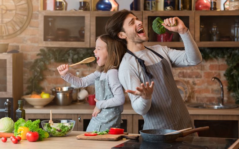 Kitchen Fun. Cheerful dad and his little daughter singing while cooking together, using spatula and broccoli as microphones