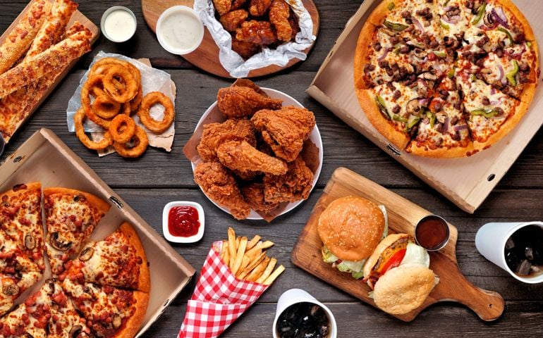 Buffet table scene of take out or delivery foods. Pizza, hamburgers, fried chicken and sides. Above view on a dark wood background.
