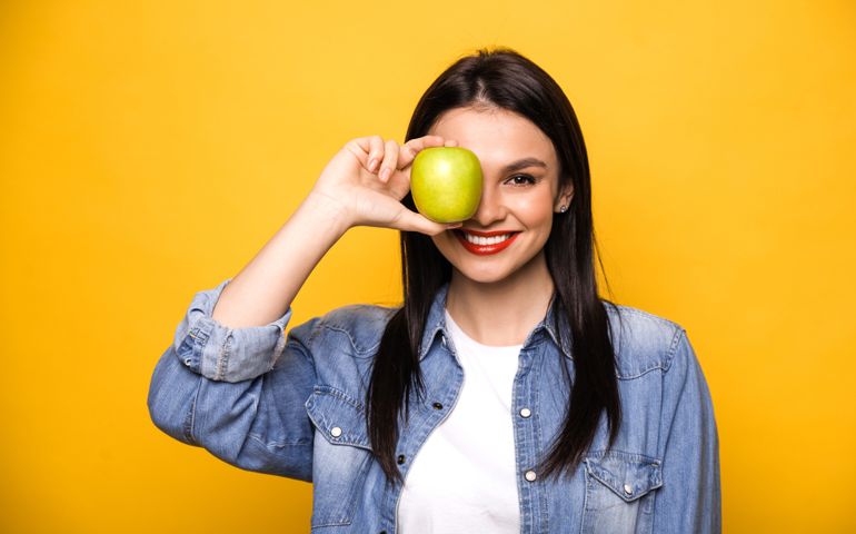 Happy caucasian woman leads a healthy lifestyle. A girl stands on an isolated orange background, holds a fresh green apple, looks into the camera covers one eye with an apple and smiles. Healthy food