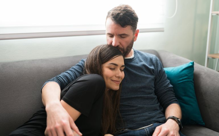 Hispanic husband kissing the top of the head of his loving beautiful wife while hugging tight on the couch during a date at home