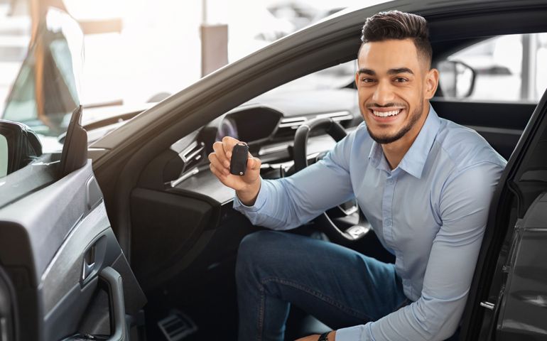 Handsome arab man customer sitting in auto and holding key, smiling at camera, buying car in showroom. Cheerful middle-eastern guy sitting inside nice sports car, happy to get new automobile