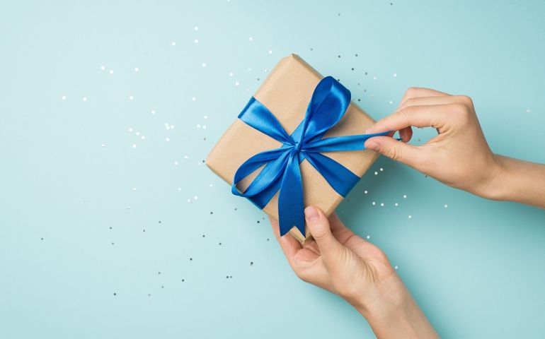 First person top view photo of hands unpacking craft paper gift box with blue satin ribbon bow over shiny sequins on isolated pastel blue background with blank space