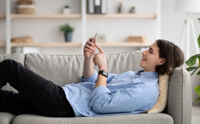 Man enjoying spare time alone lying on sofa and watching videos on cellphone, relaxing at home, using tech. Millennial guy typing on smartphone, surfing in internet in living room interior