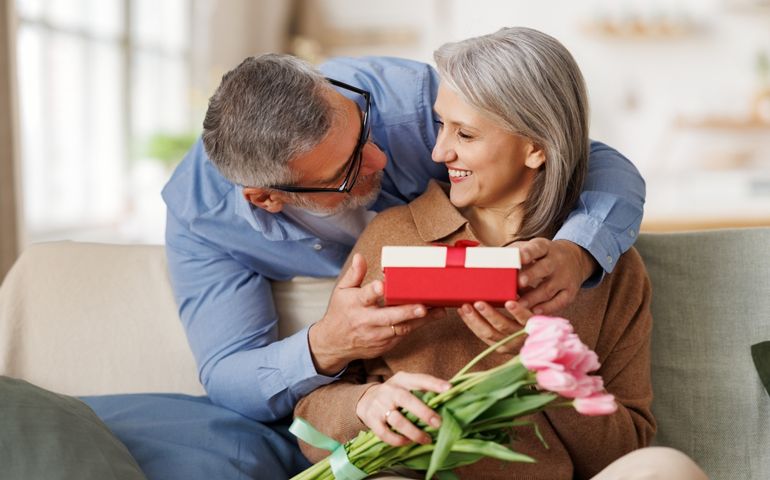 Loving senior man husband giving bouquet of tulip flowers and present gift box for Valentine's day to happy surprised wife