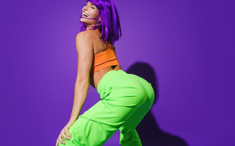Carefree woman dancer wearing colorful sportswear twerking against purple background