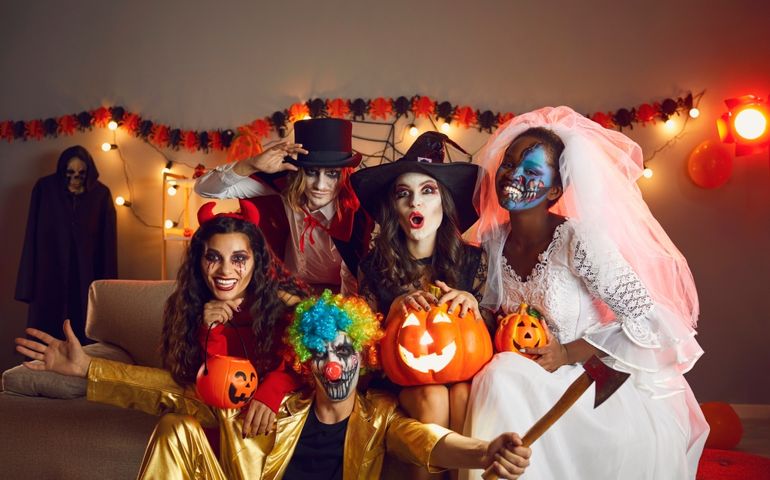 Happy adult friends in spooky costumes posing for funny group photo. Cheerful young people dressed as crazy witch, devil, vampire, dead bride and clown with axe having fun together at Halloween party