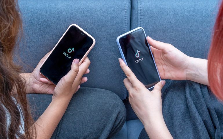 Two teenage girls with phones in their hands are sitting at home on bed. Social Media tiktok is loaded on screen.