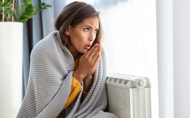 Unwell woman renter in blanket sit in cold living room hand on old radiator.suffer from lack of heat . Unhealthy young woman struggle from chill freeze at home. No heating concept.