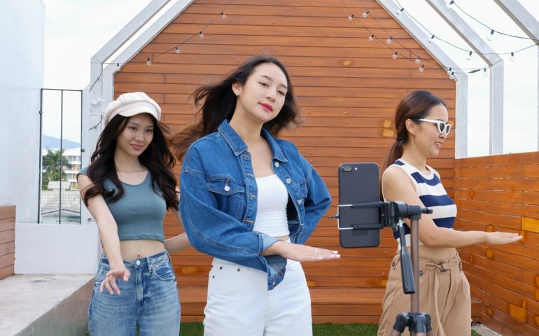 Young Asian woman with her friend tiktoker created her dancing video by smartphone camera together on rooftop outdoor at sunset. To share video to social media application