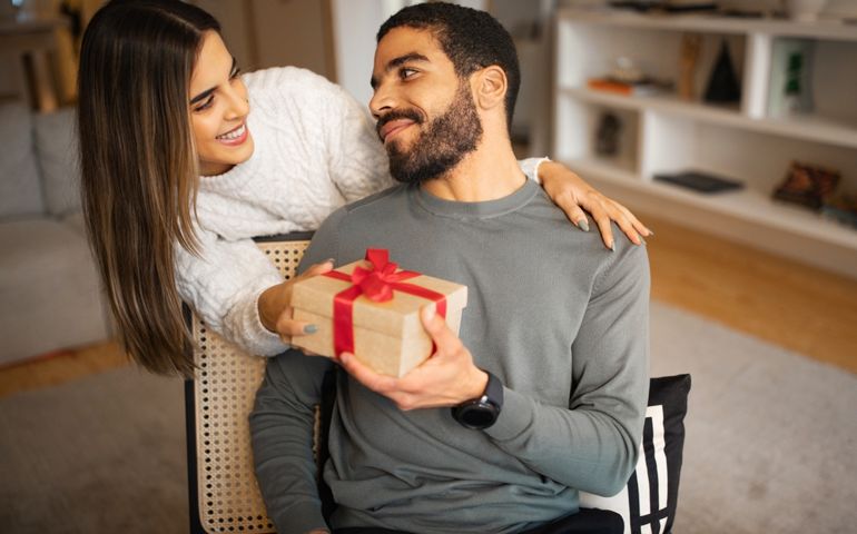 Glad young caucasian woman congratulates middle eastern male, gives box with present in room interior. Surprise for birthday, anniversary, holiday, best idea for gift, celebrate Valentine day at home