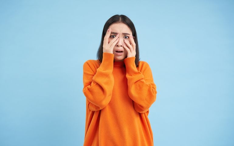 Scared Asian woman shuts her eyes and peeks through hands at smth scary and disturbing, shocked by smth horrifying, standing over blue background.