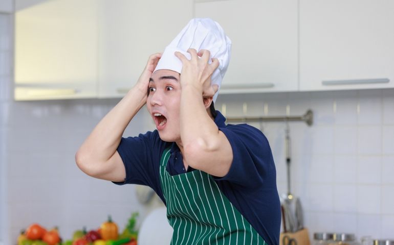 Millennial Asian excited shocked worried frightened stressed male chef wearing white tall cook hat and apron standing open mouth wide screaming shouting loud holding hands on head in home kitchen.