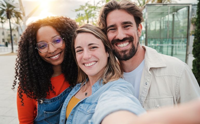 Three young adult friends smiling taking a selfie portrait and having fun together. Group of multiracial cheerful people celebrating their friendship. Two women and one man with positive expression