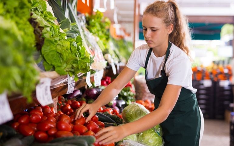 Blonde female seller in uniform working in supermarket at her first job, sorting fresh tomato