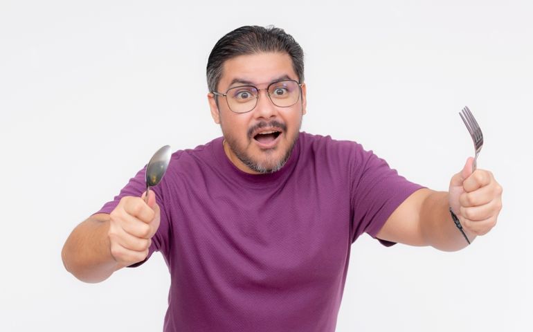 A hungry middle aged man holding a spoon and fork running towards a feast. Restaurant and other food advertisement concepts.