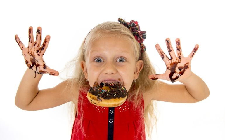 pretty little female child with long blond hair blue eyed in red dress biting donut mouth showing dirty hands with stains of chocolate syrup after eating cake smiling isolated on white background