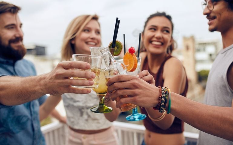 Friends having fun and drinking cocktails outdoor on a rooftop get together. Group of friends hanging out and toasting drinks outdoors. Focus on glasses.