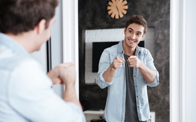 Smiling young man pointing fingers at his mirror reflection while standing at home