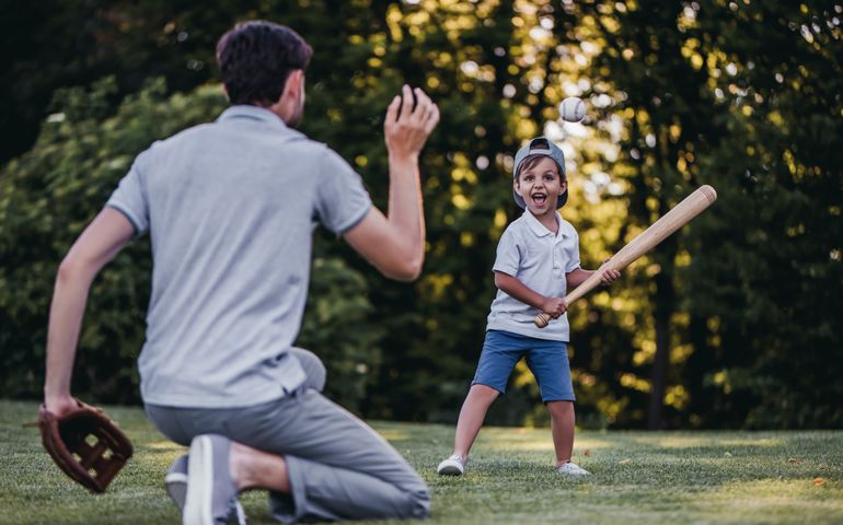 Handsome dad with his little cute sun are playing baseball on green grassy lawn