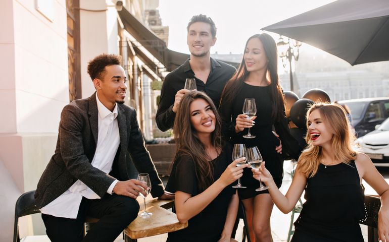 Joyful brunette man in black shirt looks away, holding glass of wine at party. Outdoor portrait of african guy in trendy jacket celebrating birthday with charming girls in cafe.