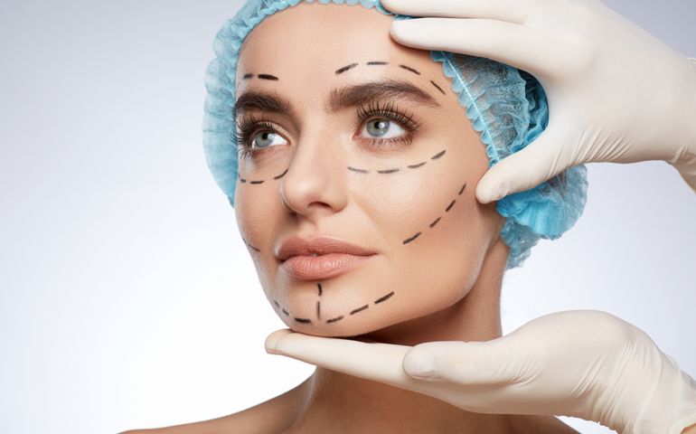 Beauty portrait of woman in blue cap looking aside, plastic surgery concept, studio. Hands in white gloves holding face of model with puncture lines, indoors