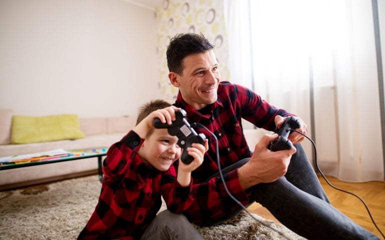 Young cheerful excited father and son in the same red shirt playing console games with gamepads while leaning against each other in a bright living room.