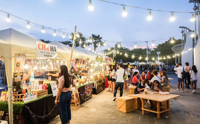 People sitting on wooden chairs,walking,eating and buying food at street food market festival in a park.